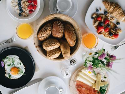 Vielseitiges Frühstück mit Brötchen, Spiegelei, Joghurt, Saft, Obst, Käse und Blumen auf weißem Tisch.