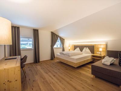 Hell eingerichtetes Schlafzimmer mit Holzboden, Doppeltbett, Sofa, Schreibtisch und großen Fenstern.