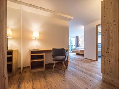 Moderner Hotelflur mit Holzfußboden, Stuhl, Tischlampe, Spiegelschrank und Blick ins helle Schlafzimmer.