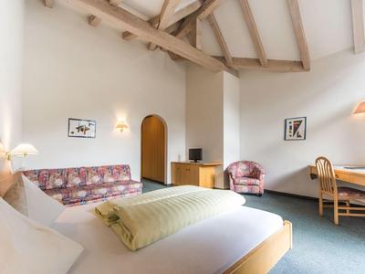 Helles Zimmer mit Holzbalken, Bett, Sofa, Schreibtisch, Stuhl und TV, Teppichboden, weiße Wände.