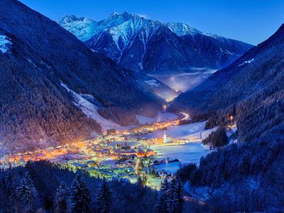 Bergdorf im Tal bei Nacht mit leuchtenden Lichtern, umgeben von schneebedeckten Bergen und blauem Himmel.