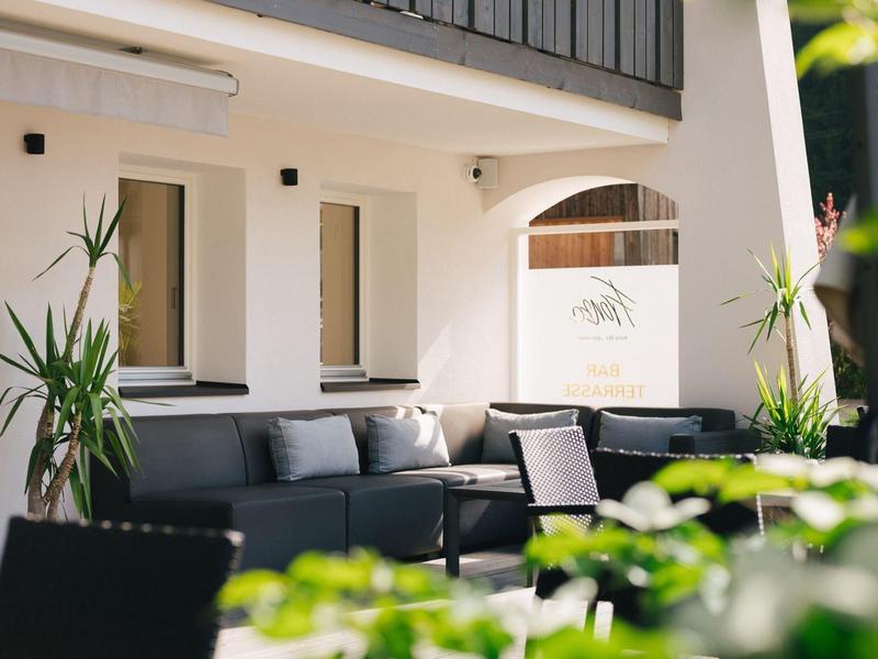 Moderner Außenbereich mit schwarzer Eckcouch, Pflanzen und sonnigem, weißem Gebäudefassade.