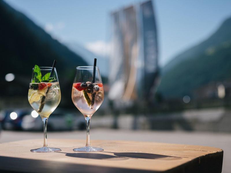 Zwei bunte Sommerdrinks in Weingläsern stehen vor Bergkulisse auf einem Holztisch.