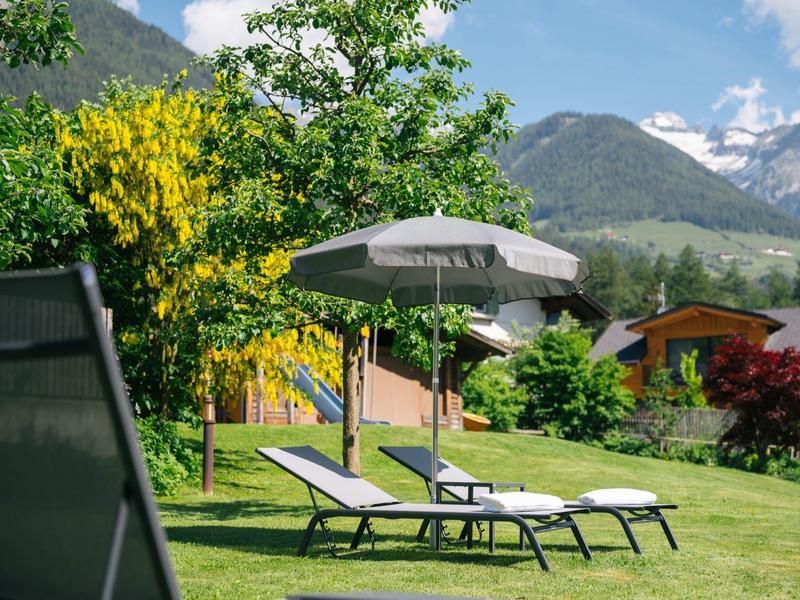 Grüner Garten mit zwei weißen Liegestühlen, grauem Sonnenschirm, Bäumen und Bergen im Hintergrund.