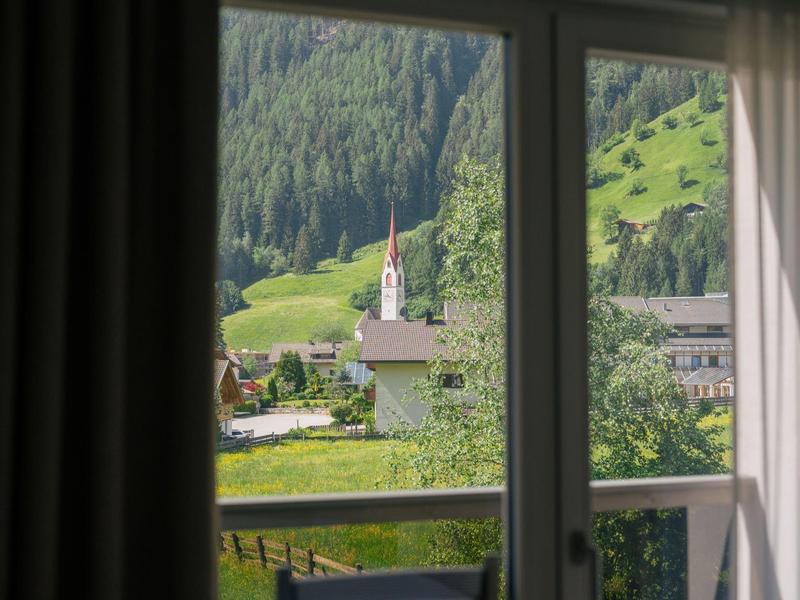Blick aus Fenster auf grünes Tal mit Kirche, Wiesen und bewaldeten Hügeln im Sonnenschein.