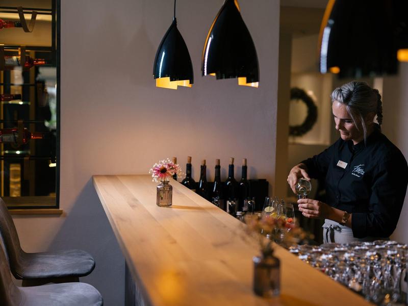Bar mit hellem Holz, schwarzen Hockern, hängenden Lampen und Barkeeper, der Flasche öffnet.