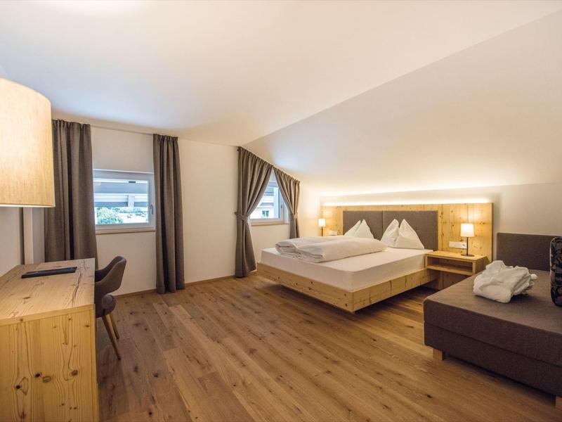 Hell eingerichtetes Schlafzimmer mit Holzboden, Doppeltbett, Sofa, Schreibtisch und großen Fenstern.