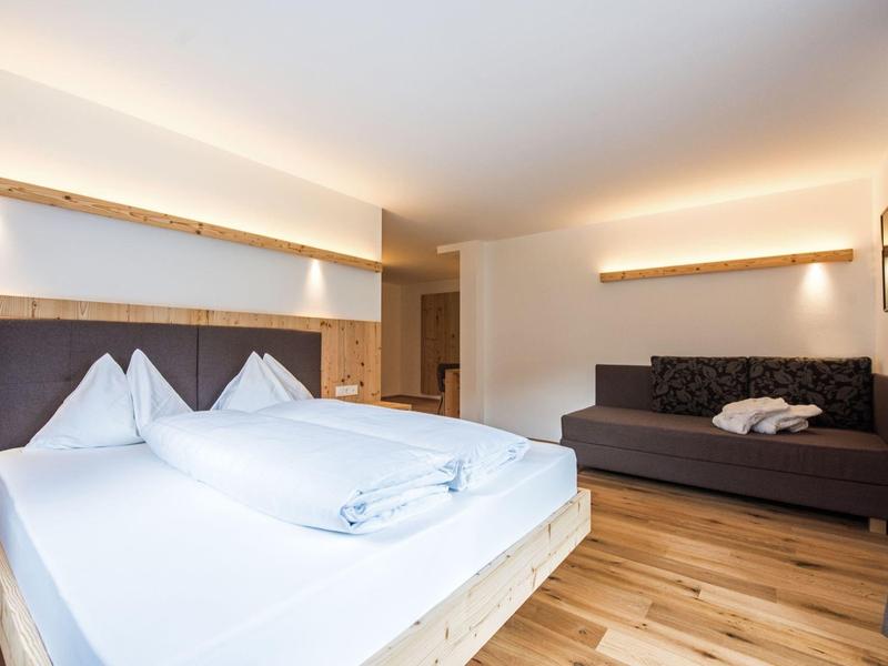 Hell eingerichtetes Schlafzimmer mit Doppelbett, Sofa, Holzboden und warmem indirektem Licht.