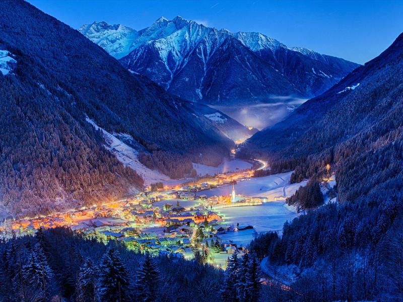 Bergdorf im Tal bei Nacht mit leuchtenden Lichtern, umgeben von schneebedeckten Bergen und blauem Himmel.