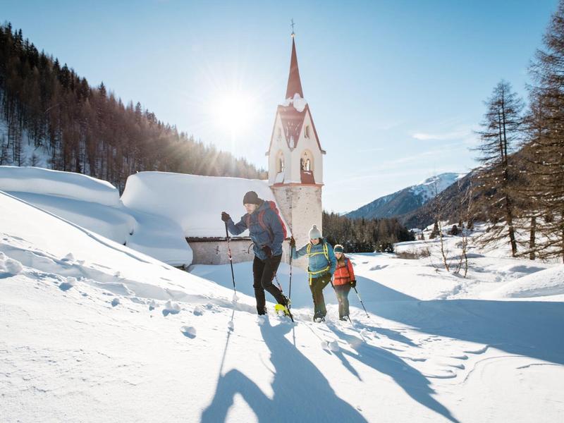 Zwei Personen wandern mit Stöcken durch verschneite Winterlandschaft vor Kirche und Bergen.