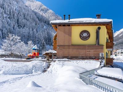 Holzhaus mit Uhr im verschneiten Winter, dahinter Wald mit schneebedeckten Bäumen und blauer Himmel.