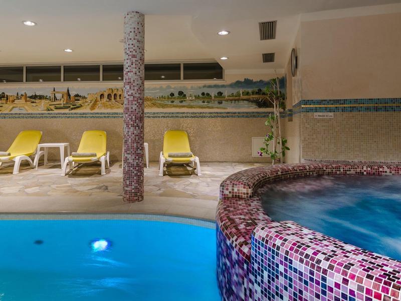 Innenpool mit blauer Wasserfläche, Whirlpool mit Mosaikfliesen und gelben Liegen im Hintergrund.