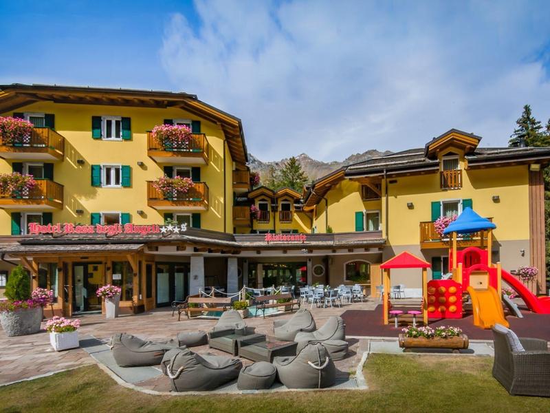 Gelbes Hotel mit Balkonblumen, Sitzbereich und buntem Kinderspielplatz bei blauem Himmel.