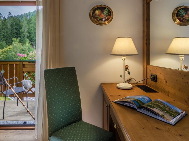 Gemütlicher Raum mit grünem Stuhl, Holztisch, Lampe und Blick auf Balkon mit Stühlen und Wald.