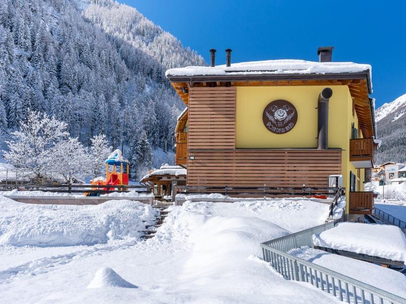Holzhaus mit Uhr im verschneiten Winter, dahinter Wald mit schneebedeckten Bäumen und blauer Himmel.