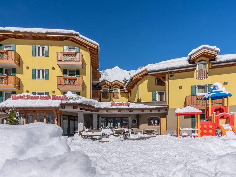 Gelbes Gebäude mit Balkon, umgeben von Schnee, darunter bunter Kinderspielplatz und blauer Himmel.