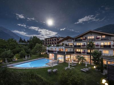 Hotelgebäude mit beleuchtetem Pool bei Nacht, umgeben von Bergen und bewölktem Himmel.