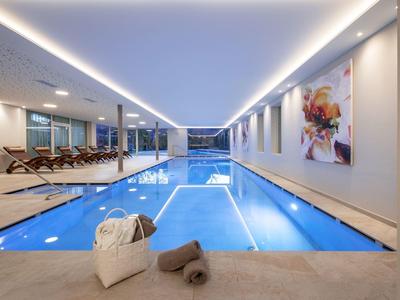 Helles Hallenbad mit blauen Fliesen, Liegestühlen, großen Bildern und weichem Licht im Raum.