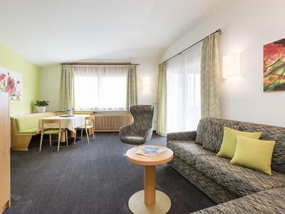 Helles Wohnzimmer mit grauem Sofa, runden Holztisch, Sessel, Esstisch und großen Fenstern mit Vorhängen.