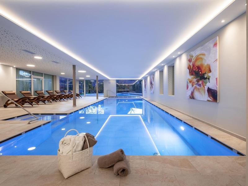 Helles Hallenbad mit blauen Fliesen, Liegestühlen, großen Bildern und weichem Licht im Raum.