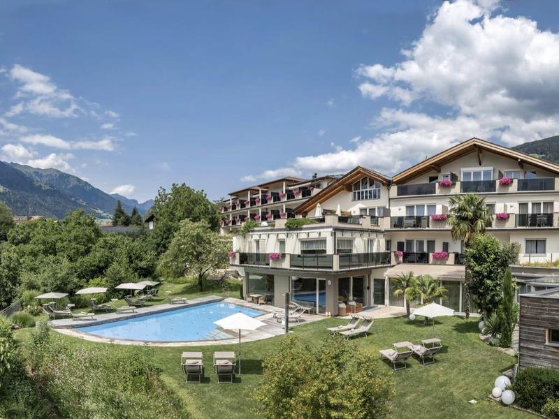 Großes Hotel mit Außenpool, Liegestühlen und Bergen im Hintergrund unter blauem Himmel.