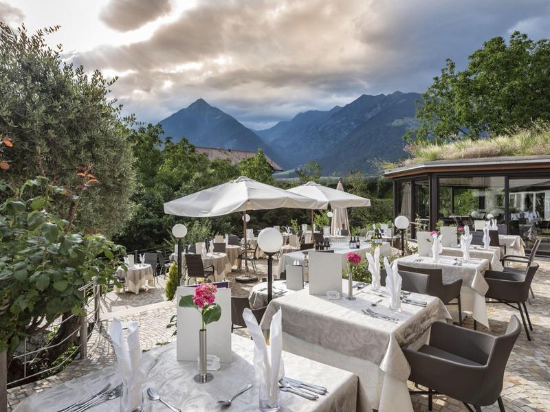 Terrassenrestaurant mit gedeckten Tischen, weißen Sonnenschirmen und Bergblick bei bewölktem Himmel.