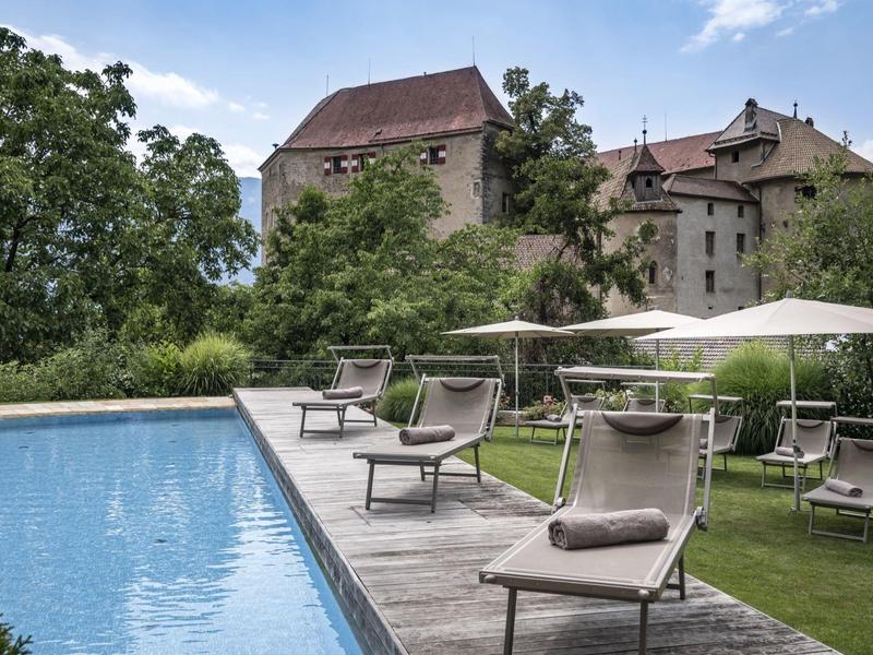 Moderner Pool mit Liegestühlen und Sonnenschirmen vor mittelalterlicher Burg in grüner Umgebung.