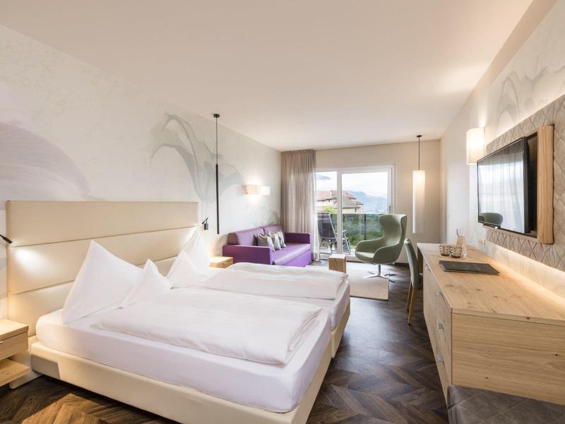 Helles Schlafzimmer mit großem Bett, Holzmöbeln, Sitzbereich und Balkon mit Blick ins Grüne.