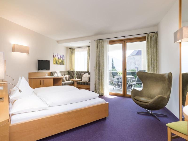 Modernes Hotelzimmer mit Doppelbett, Fernsehgerät, Balkon und bequemen Sessel in warmen Farbtönen.