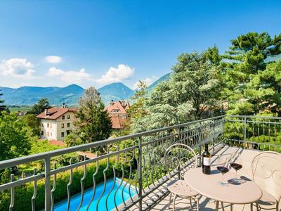 Balkon mit Metallgeländer, kleinem runden Tisch, zwei Stühlen, Blick auf Bäume, Häuser und Berge.