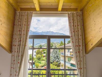 Blick durch ein großes Fenster mit Gardinen auf Berge, grünes Tal und Häuser bei klarem Himmel.
