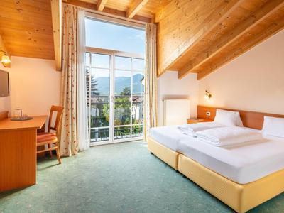 Gemütliches Schlafzimmer mit Holzbalkendecke, großem Fenster, Doppelbett, Schreibtisch und grüner Teppichboden.