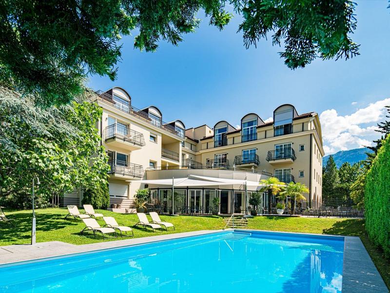 Hotel mit Außenpool, Liegestühlen und grüner Gartenlandschaft unter blauem Himmel.