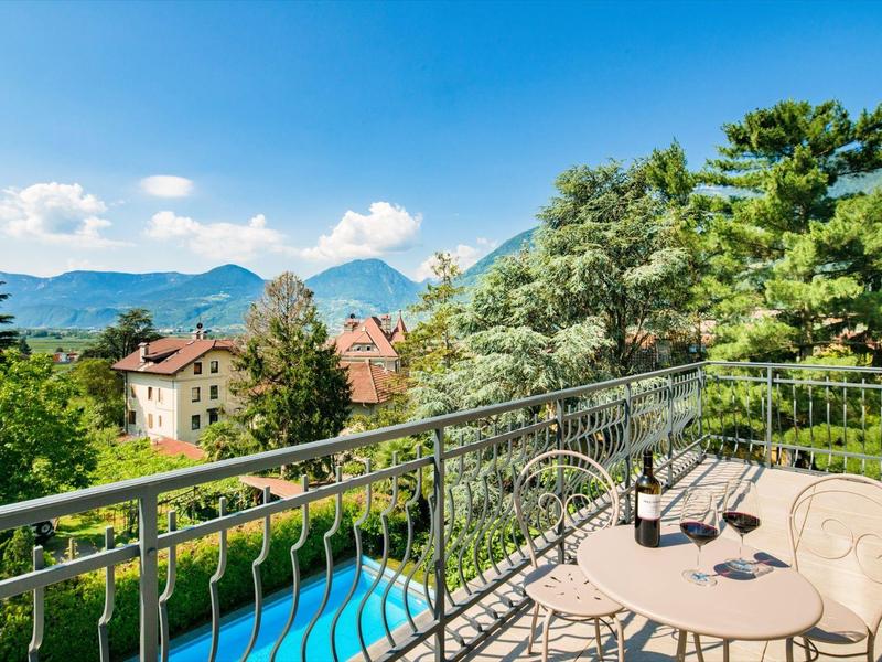 Balkon mit Metallgeländer, kleinem runden Tisch, zwei Stühlen, Blick auf Bäume, Häuser und Berge.