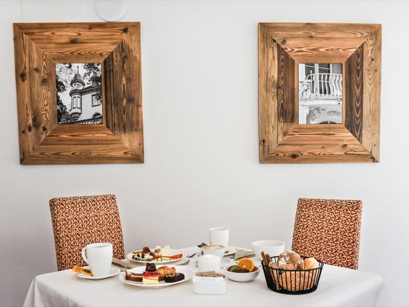 Ein gedeckter Tisch mit Kuchen, Tassen, Besteck und einem Brotkorb vor zwei Holzbilderrahmen an weißer Wand.