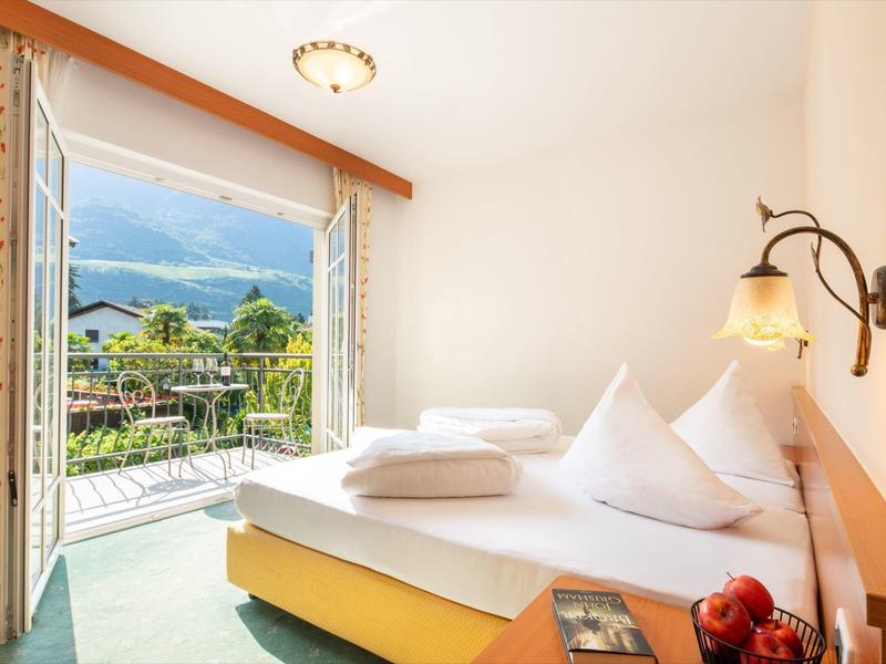 Hell eingerichtetes Schlafzimmer mit großem Bett, offenem Balkon und Blick auf grüne Berge.