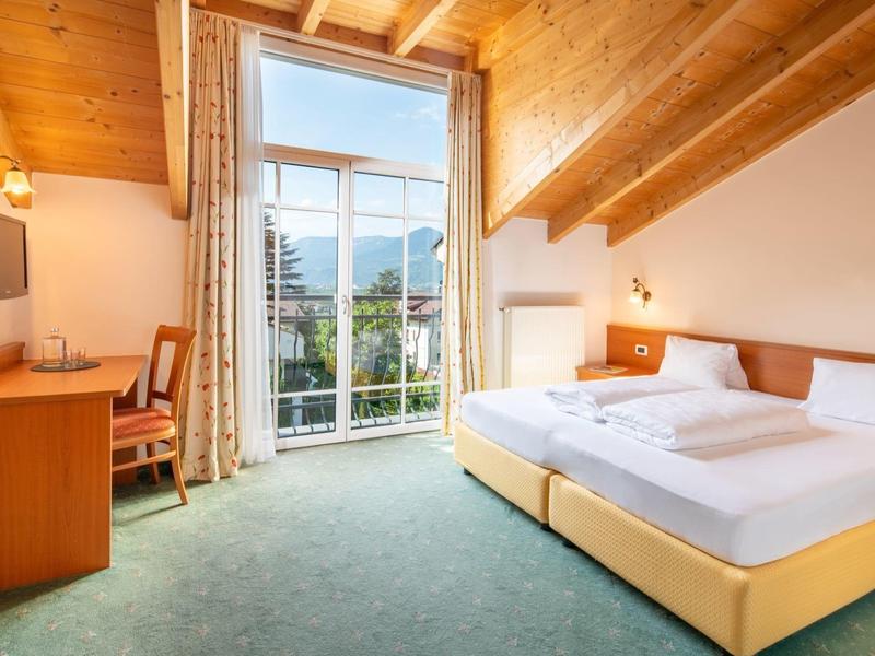 Gemütliches Schlafzimmer mit Holzbalkendecke, großem Fenster, Doppelbett, Schreibtisch und grüner Teppichboden.