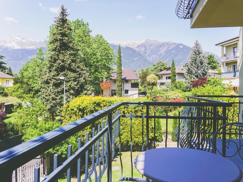 Balkon mit blauem Tisch und Stuhl, grüne Bäume, Häuser und Berge im Hintergrund unter blauem Himmel.