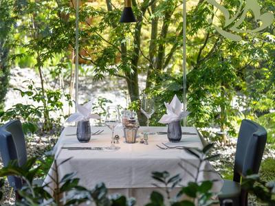 Tisch mit weißer Decke und zwei gedeckten Plätzen, umgeben von grünem Laub, nahe Wasser.