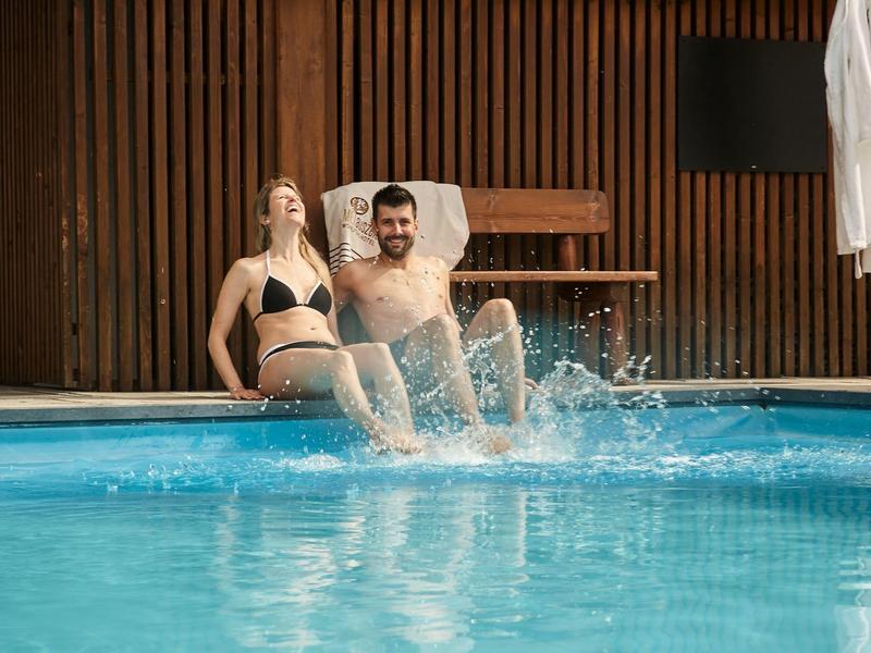 Ein Mann und eine Frau sitzen am Beckenrand; der Mann plantscht mit den Füßen im Wasser.
