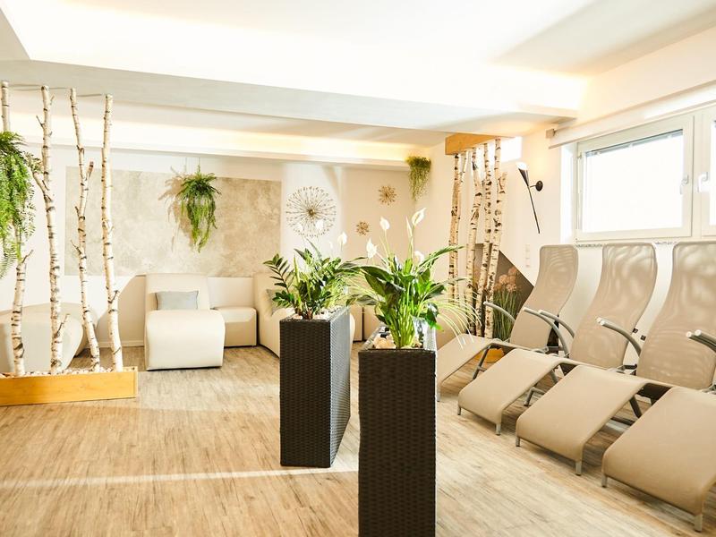 Heller Wellnessraum mit bequemen Liegen, weißen Holzdetails und grünen Pflanzen in modernen Pflanzgefäßen.