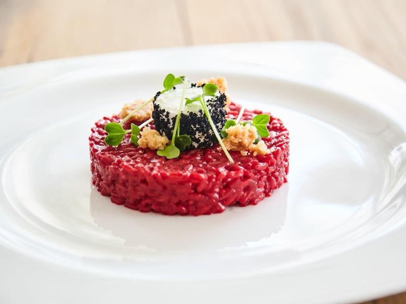 Rund geformtes rotes Risotto auf weißem Teller, garniert mit grünen Kräutern und dunklen Beeren.