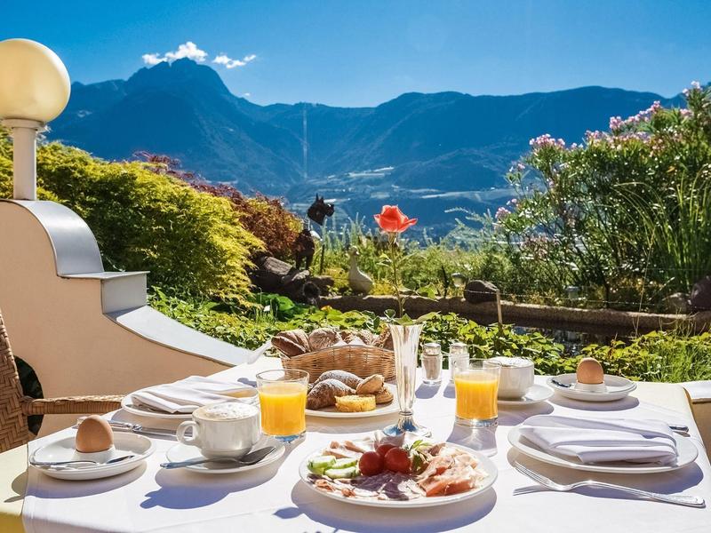 Tisch mit Frühstück und Getränken auf Terrasse mit Bergblick und blauem Himmel im Hintergrund.