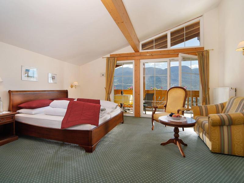 Helles Hotelzimmer mit Doppelbett, Sessel, Sofa, Holzmöbeln und Balkon mit Bergblick.