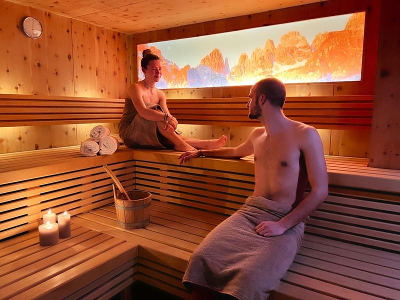 Zwei Personen entspannen in einer Sauna aus hellem Holz mit Kerzenlicht und Berglandschaftbild.
