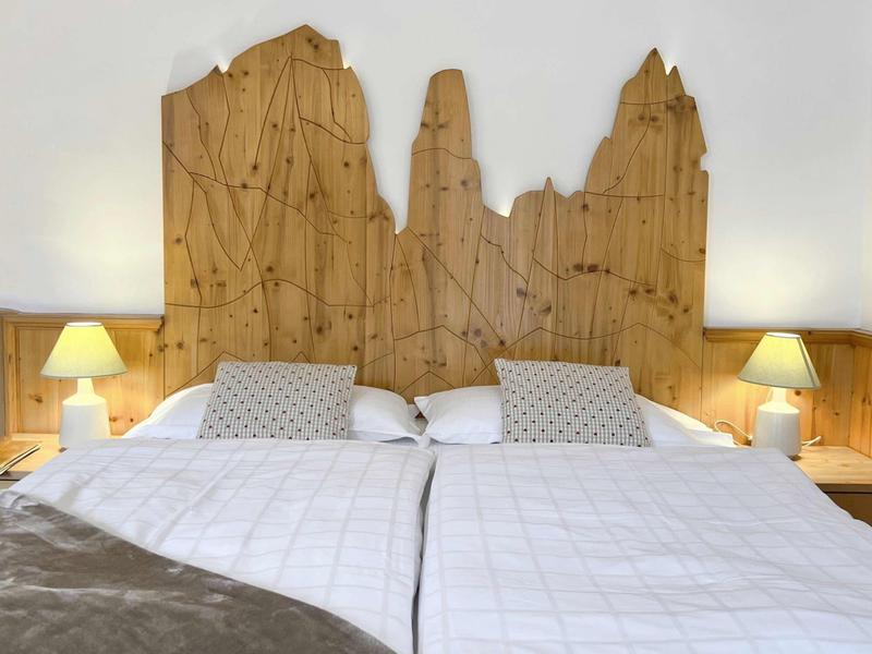 Zwei Einzelbetten mit weißer Bettwäsche, Holz-Vertäfelung und "Baum"-förmigem Kopfteil, je mit Lampe.