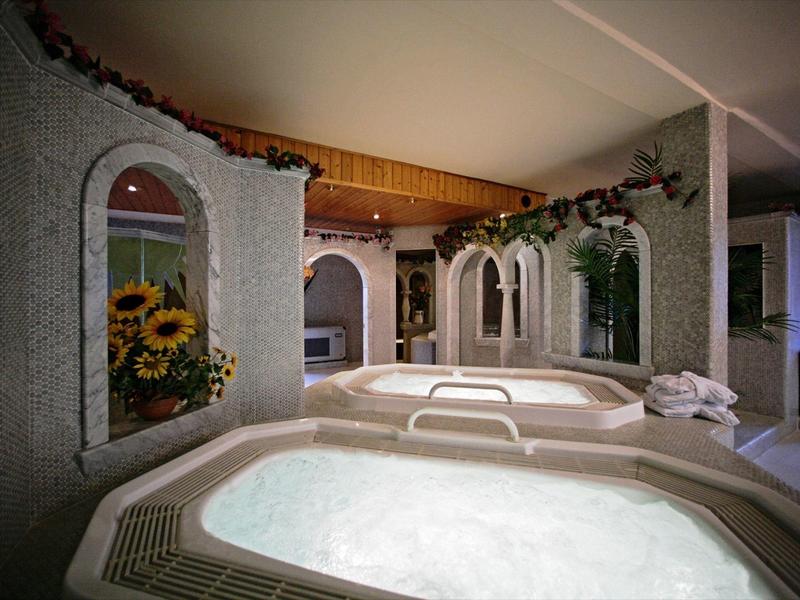 Spa lumineux avec jacuzzi, fleurs, fenêtres voûtées et plafond à poutres en bois dans un cadre paisible.