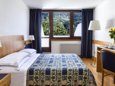 Habitación de hotel luminosa con cama doble, escritorio, silla, ventanas con vista a zonas verdes y cortinas