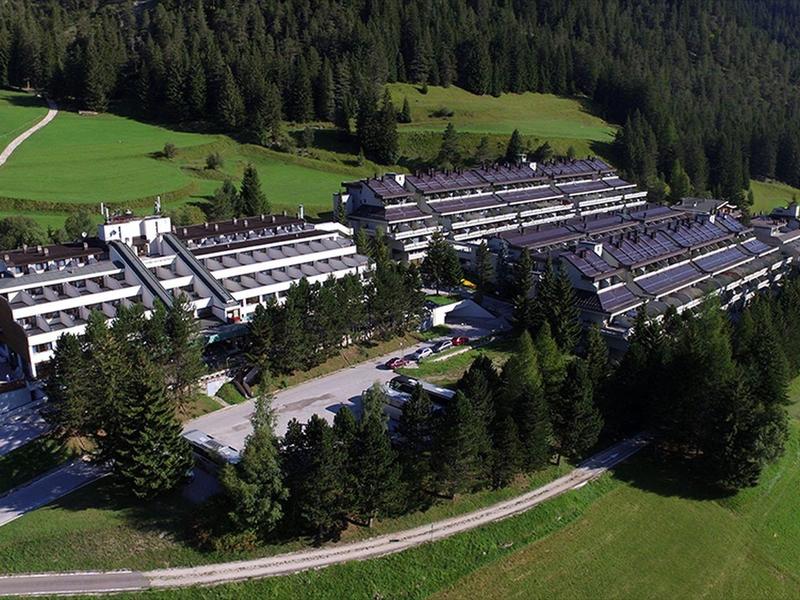 Großes Hotelgebäude umgeben von Wiesen und Wald in einer grünen Landschaft.