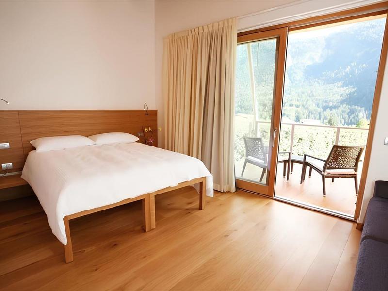 Camera d'hotel luminosa con letto matrimoniale, pavimento in legno e balcone con vista sulle montagne.
